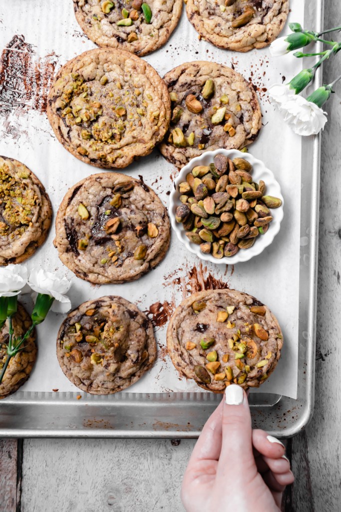 flat lay of the pistachio dark chocolate chunk cookies on a pub table backdrop, with a hand picking up one of the cookies