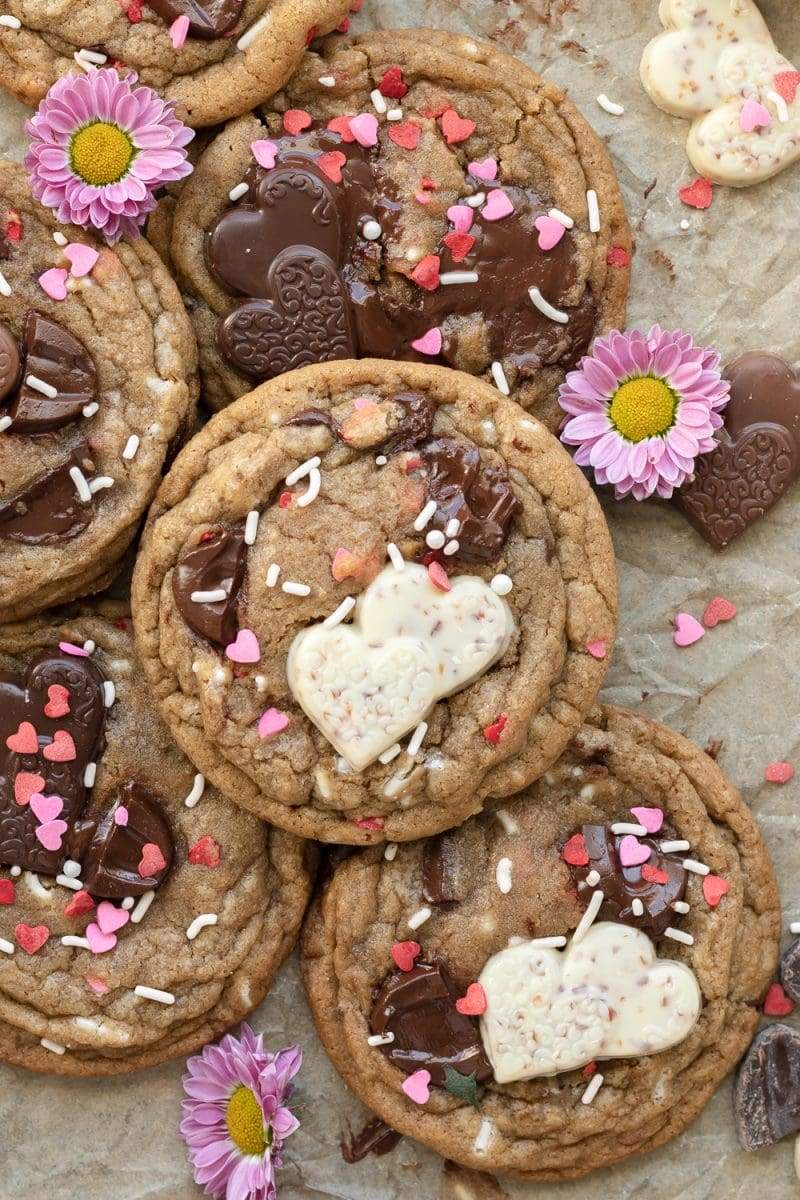 Flat lay of the valentine's day chocolate chip cookies stacked on top of each other, surrounded by chocolate hearts and purple flowers