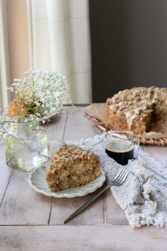 A piece of pistachio coffee cake on a plate with the whole cake in the background.